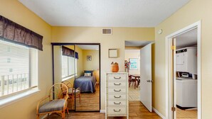 A room with a bed, a dresser, a chair, a dining table, and a laundry area. - Beachfront condo with balcony overlooking Gulf of Mexico & resort pool (Gulf Shores)