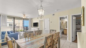 A dining area with a wooden table and chairs, a ceiling fan, and a television. - Beachfront condo with balcony overlooking Gulf of Mexico & resort pool (Gulf Shores)