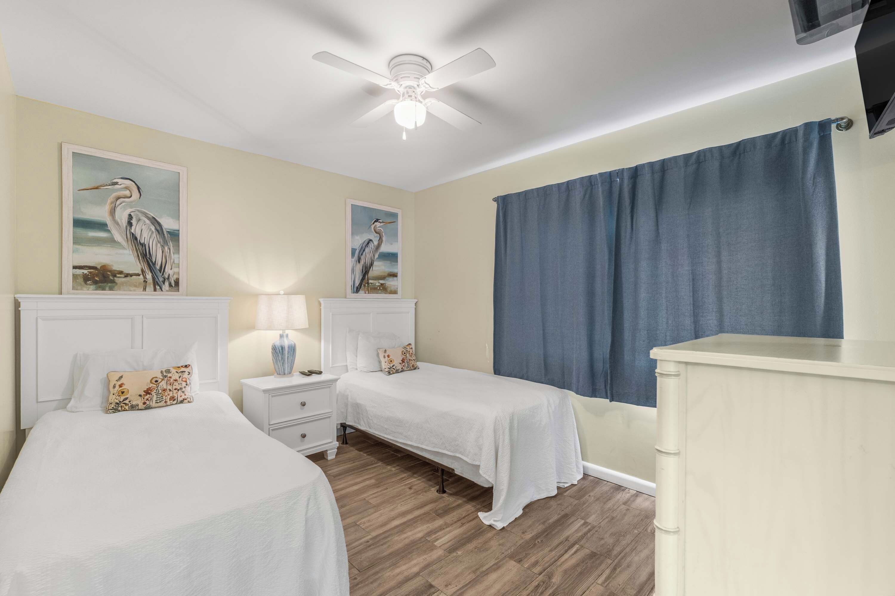 A hotel room with two beds, a ceiling fan, a nightstand with a lamp, and a large window with a blue curtain.