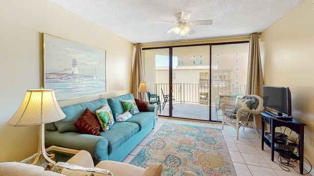 A living room with a sofa, a television, a ceiling fan, and a view of the outside balcony. - Beachfront condo with balcony overlooking Gulf of Mexico & resort pool (Gulf Shores)