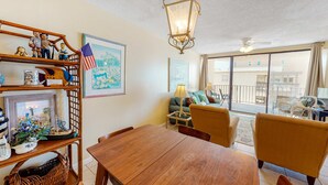A dining area with a wooden table and chairs, a hanging light fixture, and a view of the outside through large windows. - Beachfront condo with balcony overlooking Gulf of Mexico & resort pool (Gulf Shores)