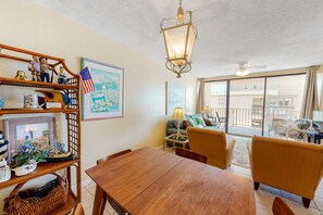Un espace repas avec une table et des chaises en bois, un luminaire suspendu et une vue sur l’extérieur grâce à de grandes fenêtres.