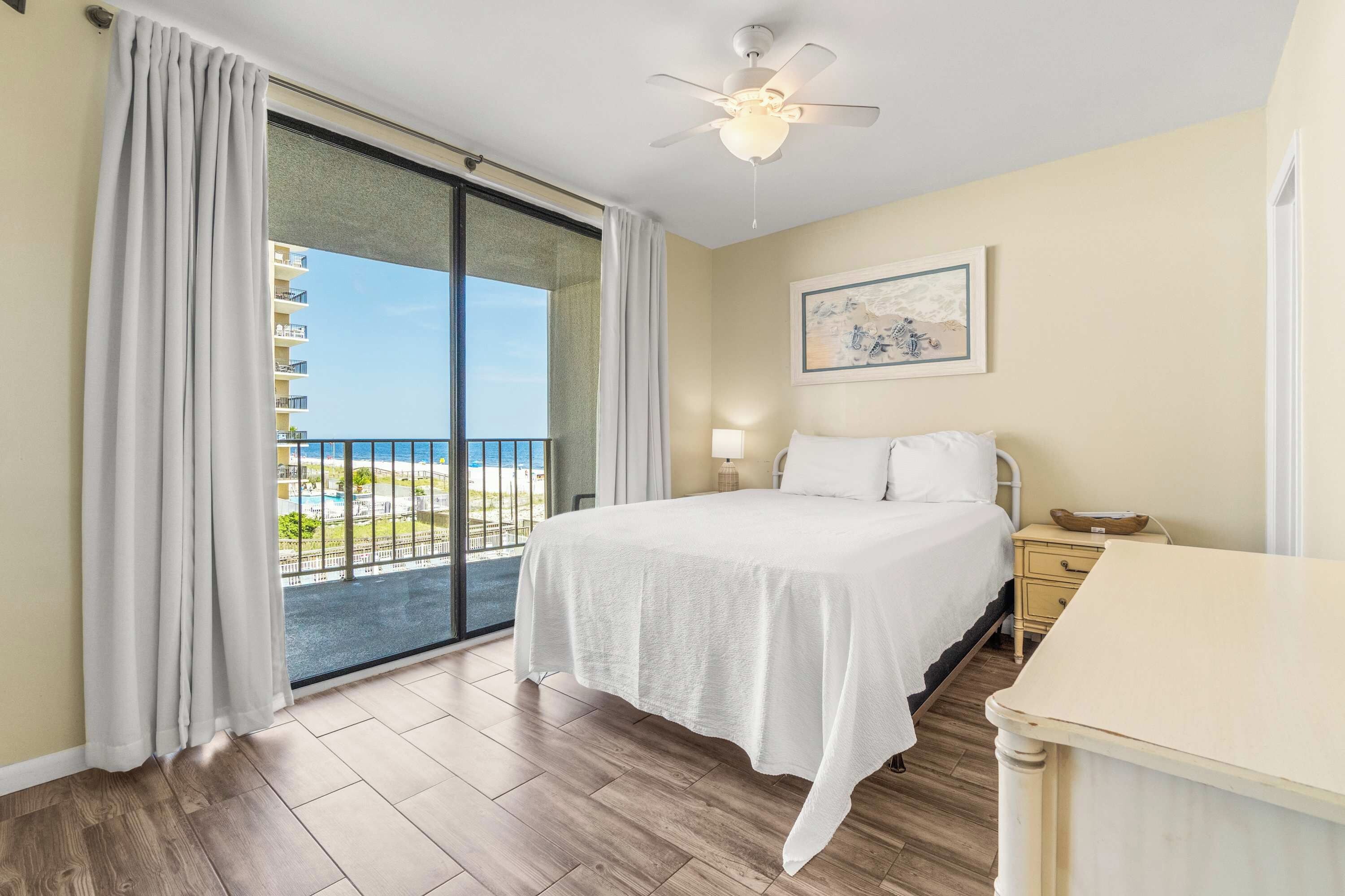 Ein Hotelzimmer mit einem Bett, einem Nachttisch, einer Lampe, einem Deckenventilator und Blick auf den Strand durch eine Schiebetür.