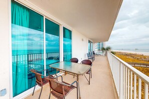 Un balcon avec une table et des chaises, de grandes fenêtres avec des rideaux et une vue sur la plage et l’océan.