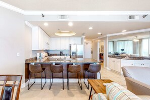 A modern kitchen with white cabinets, a central island with bar stools, and a dining area with a wooden table and striped sofa. - Gulf-front condo w/ shared beachside pools, hot tubs Near top attractions (Gulf Shores)