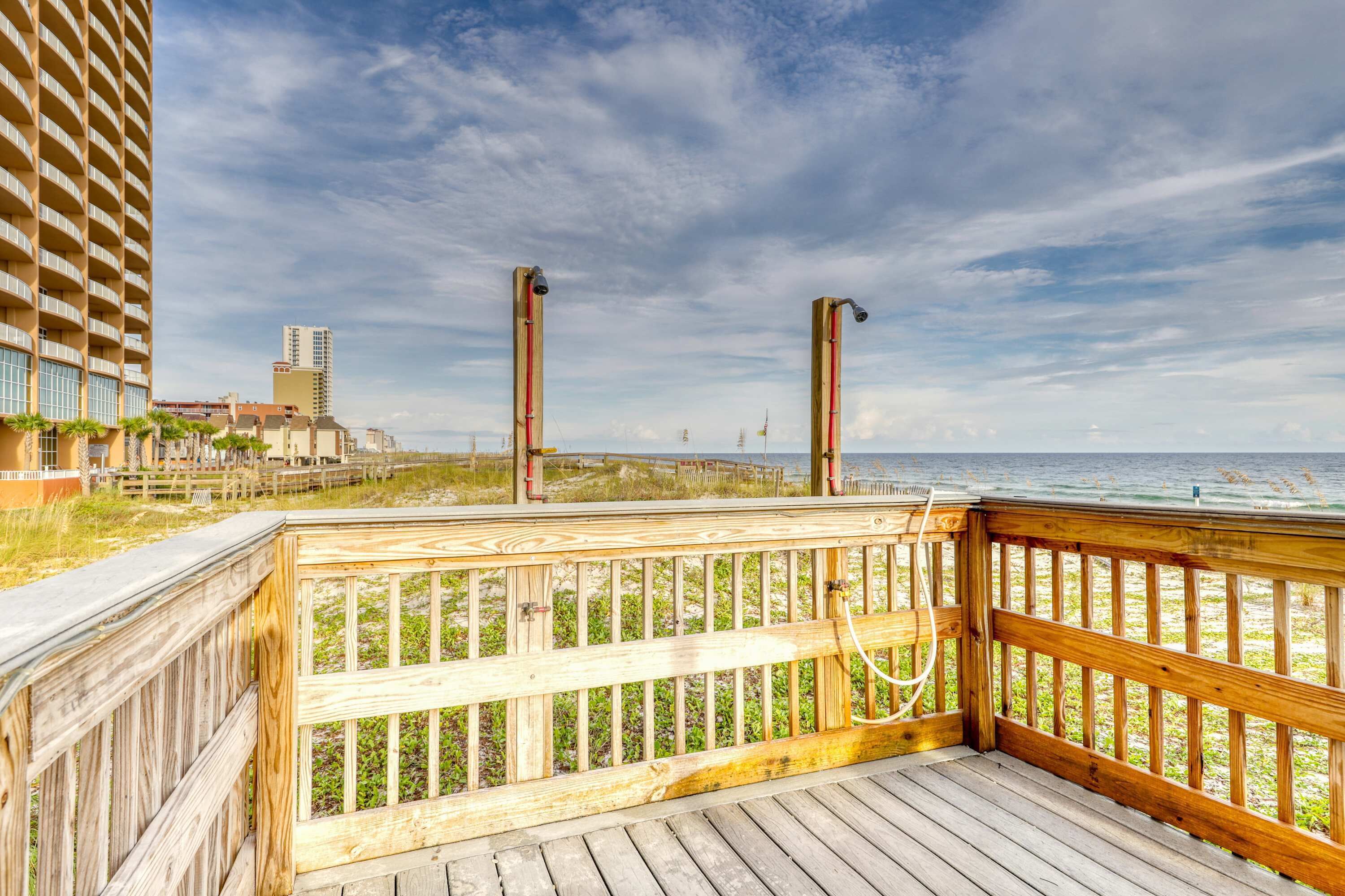 A wooden deck with a railing overlooking a beach, a tall building to the left, and a clear sky.
