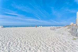 Ein Sandstrand mit blauem Himmel, einer Umzäunung und Gebäuden im Hintergrund.