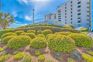 Ein angelegter Garten mit ordentlich geschnittenen Büschen und ein Gebäude im Hintergrund, auf dem 'Island Winds East' steht.