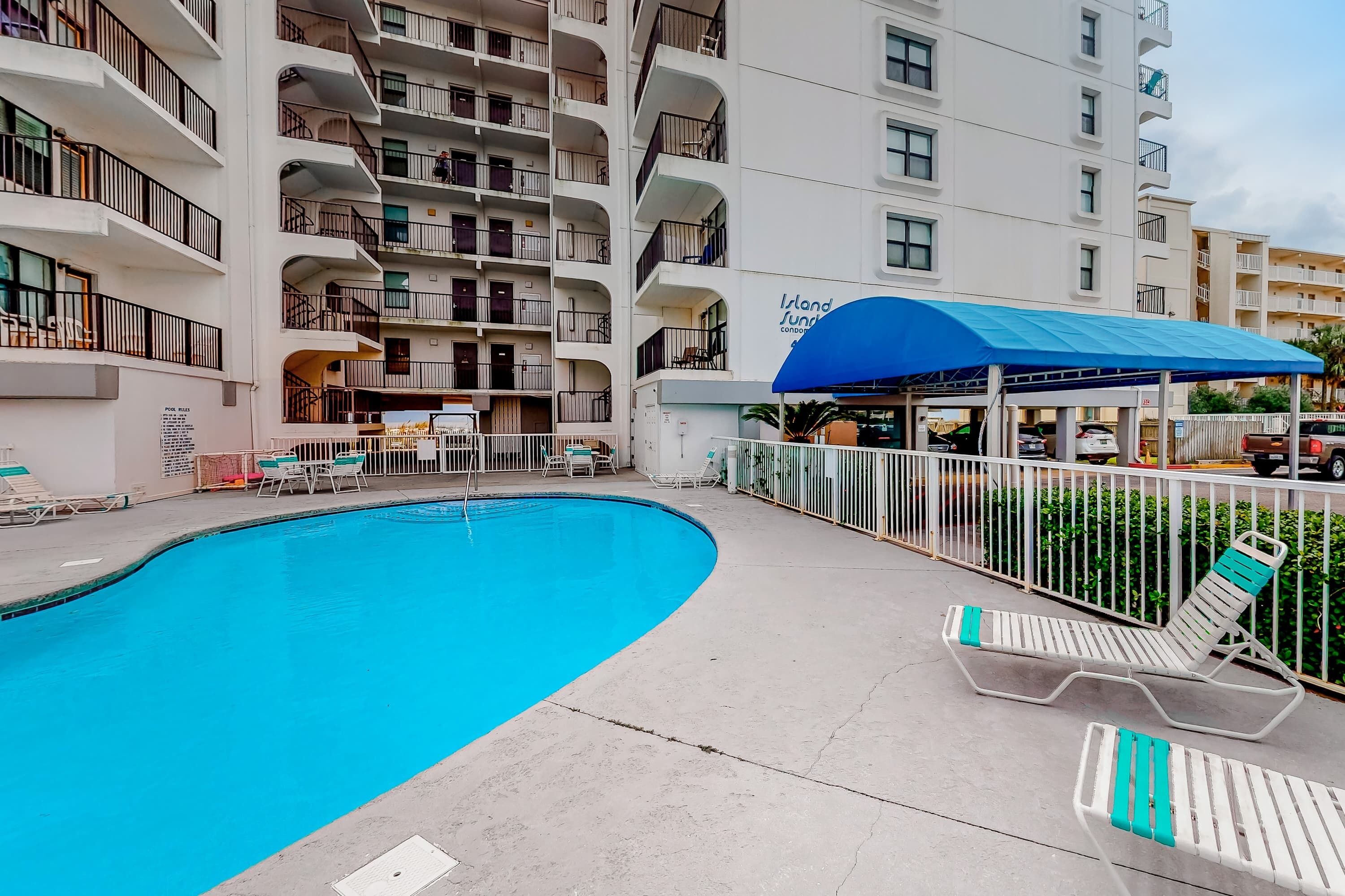 A swimming pool with lounge chairs, a covered seating area, and a multi-story building with balconies.