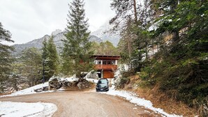 Una cabaña con camino de grava, rodeada de terreno y árboles cubiertos de nieve.