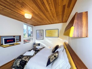 Een slaapkamer met een houten plafond, een bed met witte lakens, een tv aan de muur waarop 'NETFLIX' staat en twee kunstwerken in lijsten.