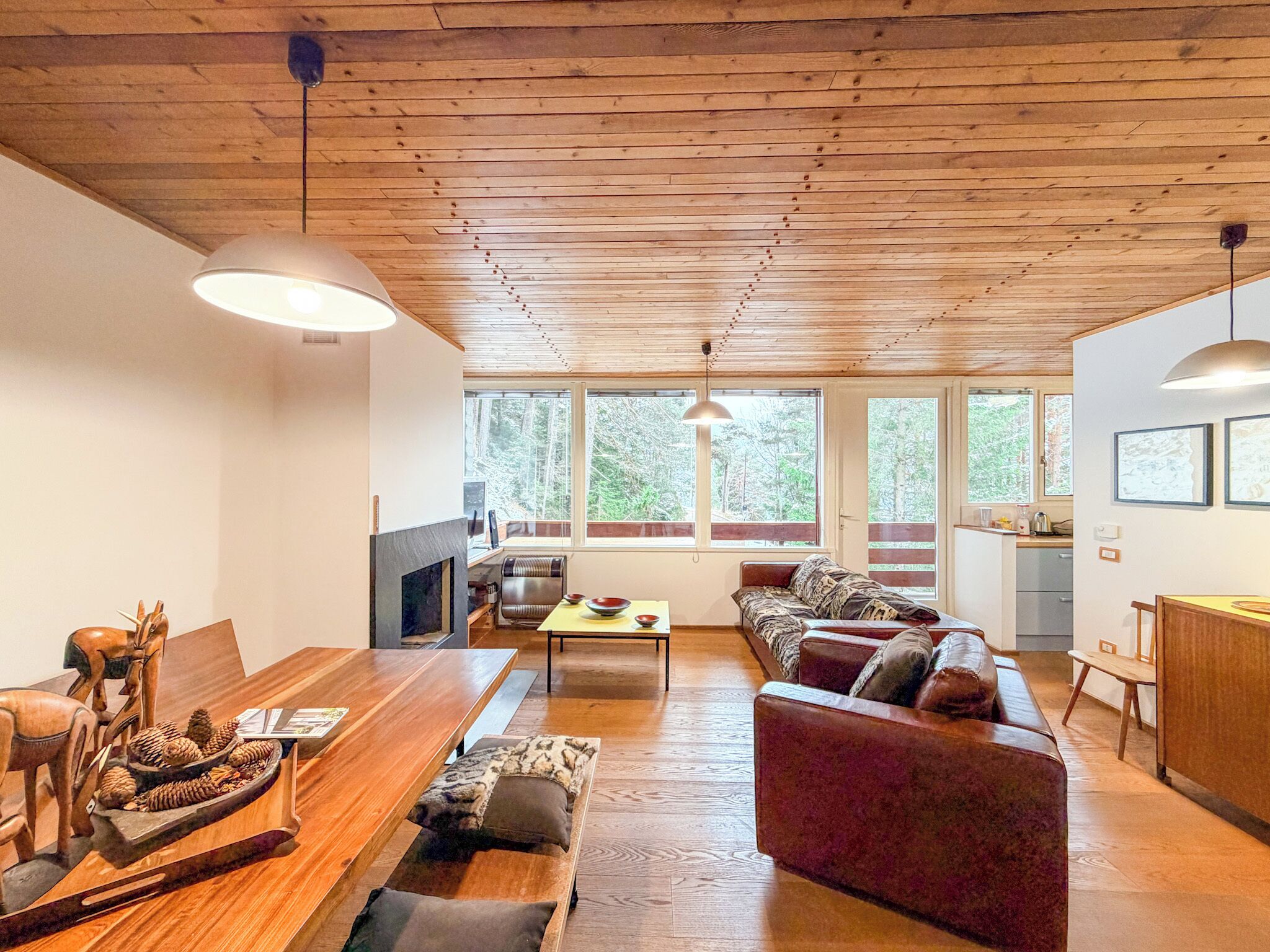 Amplia sala de estar con techo de madera, chimenea, sofá, mesa de comedor y grandes ventanales con vistas al bosque.