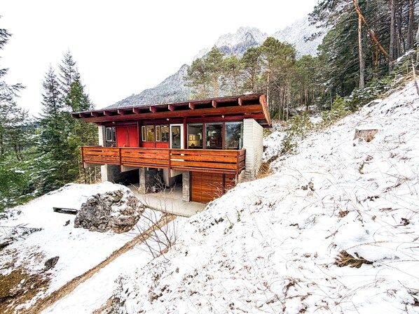 Een moderne houten blokhut met een terras, gebouwd in een heuvel met uitzicht op besneeuwde bergen.