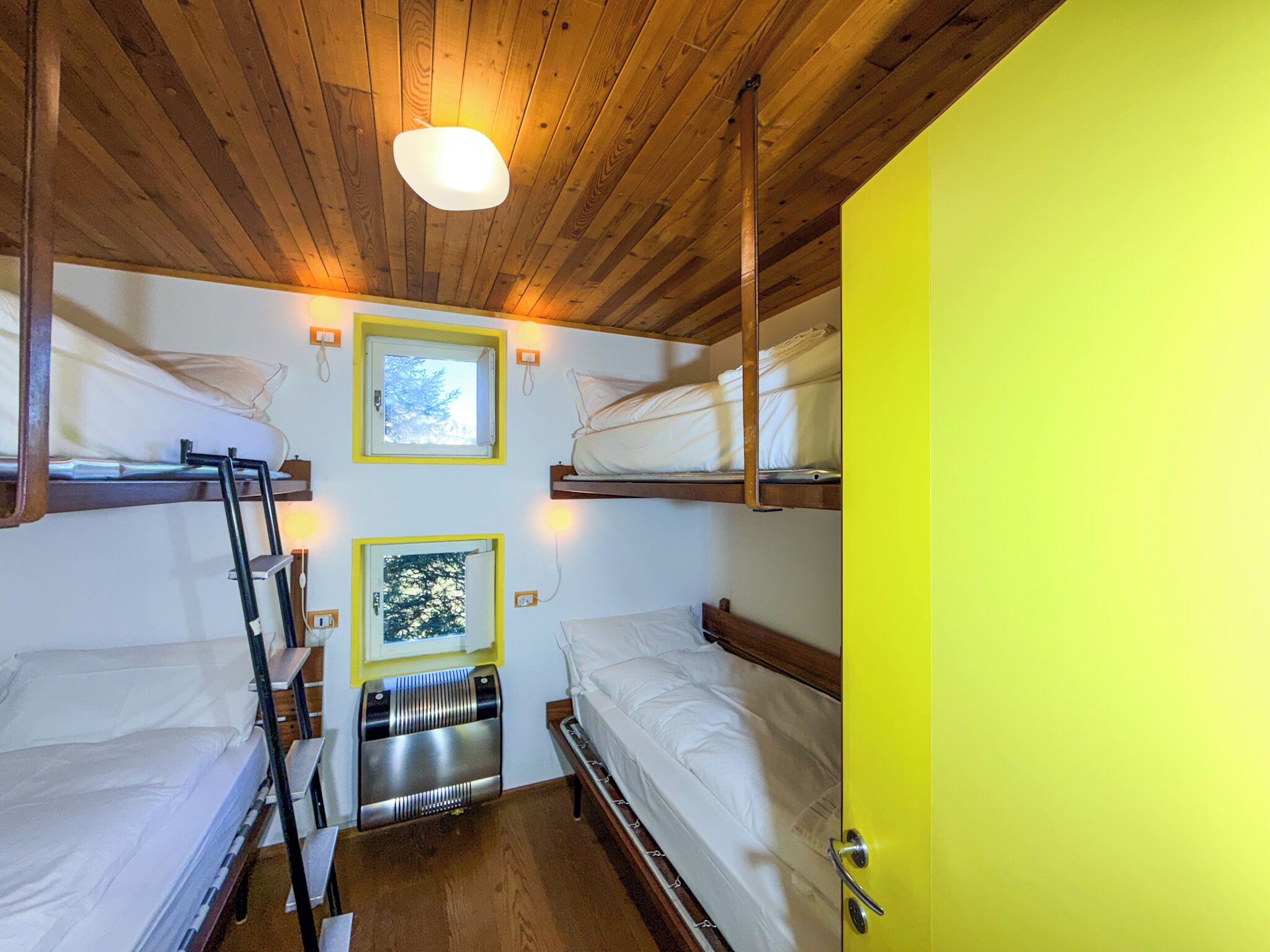 A room with bunk beds, a ladder, a radiator, and a window with a view of trees.