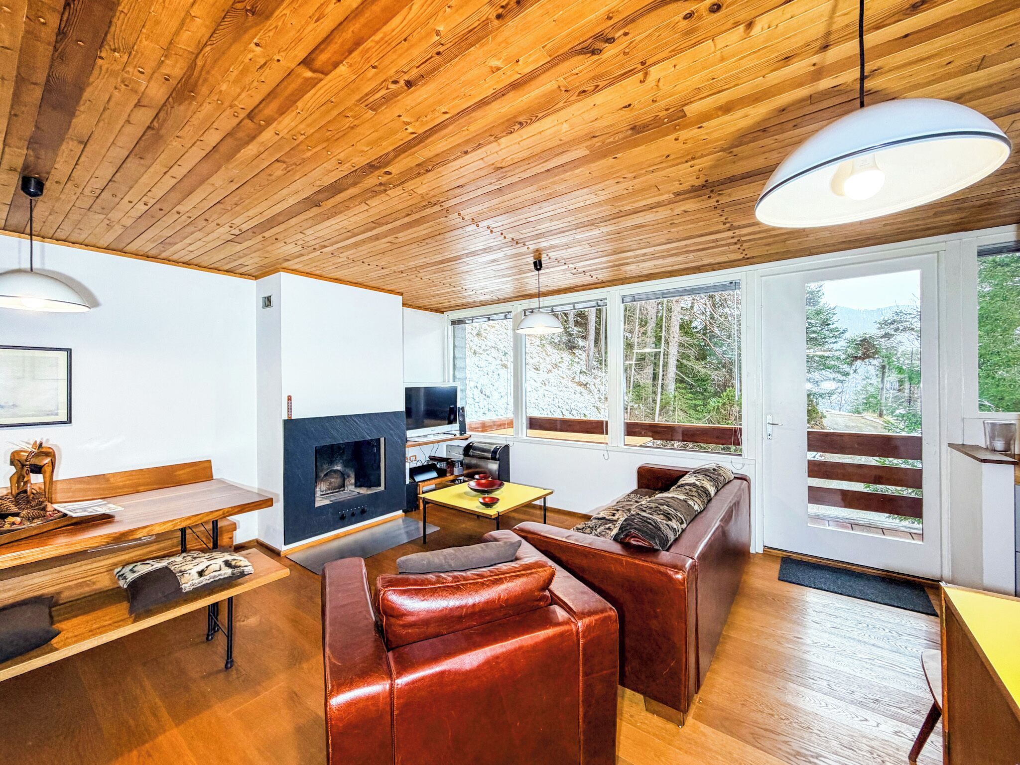 Interior acogedor de una cabaña con techo de madera, chimenea, sofá, mesa de comedor y ventanas amplias con vistas al bosque.