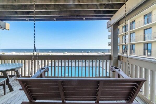 A wooden swing chair overlooking a pool and beach. - Ocean-view condo with pool access, outdoor seating, central AC & WiFi (Gulf Shores)