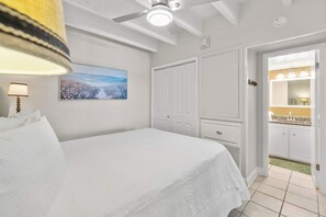 A bedroom with a bed, a ceiling fan, a painting, a door leading to a bathroom, and a tiled floor. - Ocean-view condo with pool access, outdoor seating, central AC & WiFi (Gulf Shores)
