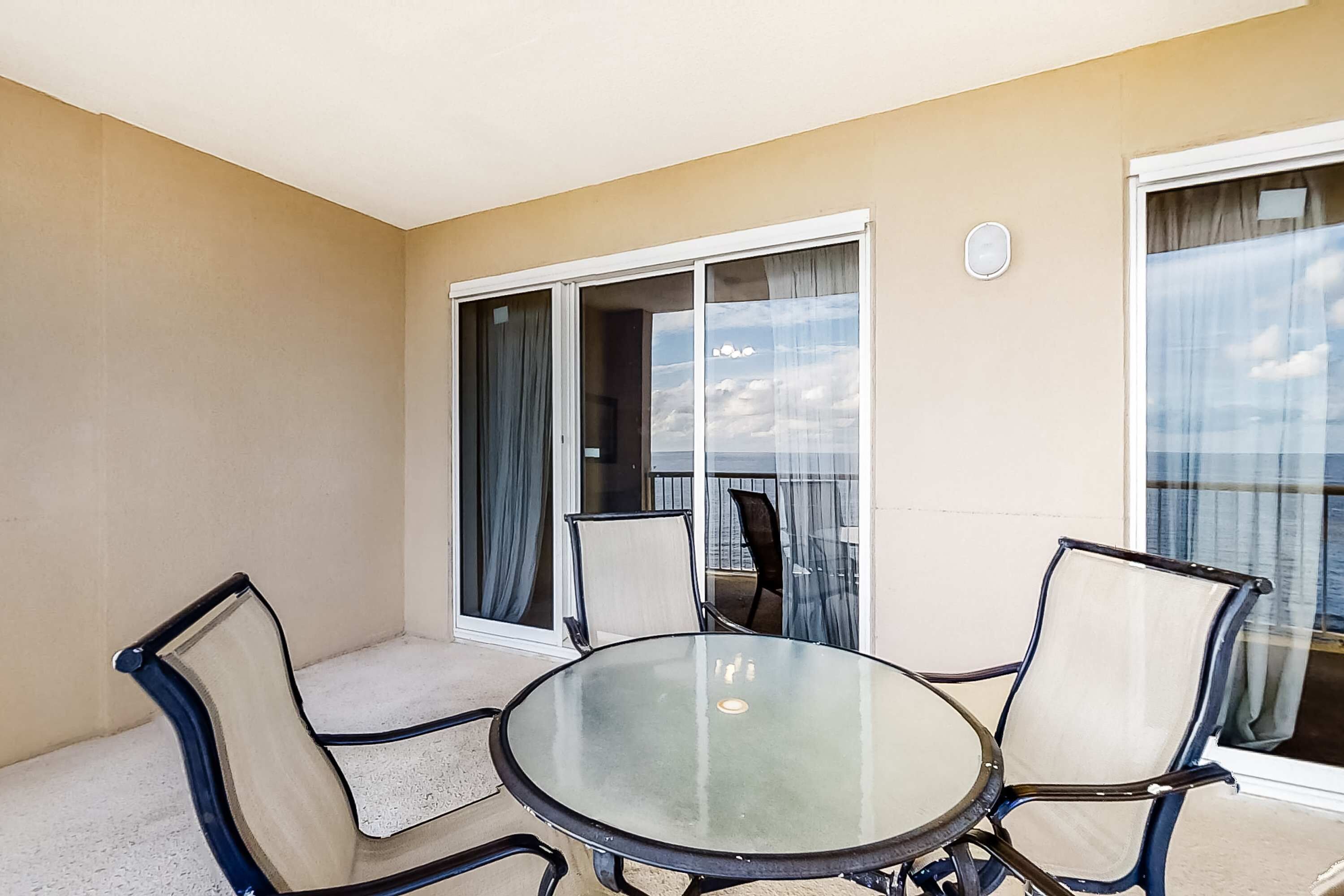En balkon med et glasbord og stole, en skydedør i glas samt udsigt til himlen.