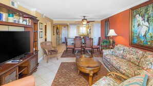 A living room with a TV, wooden furniture, a dining area, and a painting on the wall. - Beachfront Condo with Shared Outdoor Pool, Central AC, Private Balcony, Views, WiFi (Gulf Shores)