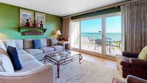 A living room with a sofa, armchair, and a glass coffee table. There is a balcony with a view of the sea. - Gulf-Front Condo with Shared Outdoor Pool, Private Balcony, Incredible Views & WiFi (Gulf Shores)