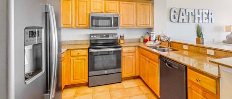 A modern kitchen with wooden cabinets, stainless steel appliances, and a 'GATHER' sign on the wall.