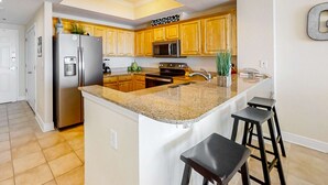 A kitchen with wooden cabinets, a stainless steel refrigerator, a microwave, a stove, and a granite countertop. - Beachfront 5th-floor condo with balcony, AC, pool, hot tub & view (Gulf Shores)