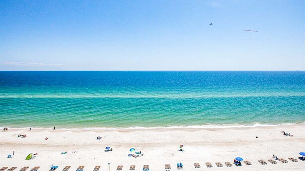 A beach with clear blue water, a sandy shore, and a few umbrellas and lounge chairs. - Beachfront 5th-floor condo with balcony, AC, pool, hot tub & view (Gulf Shores)