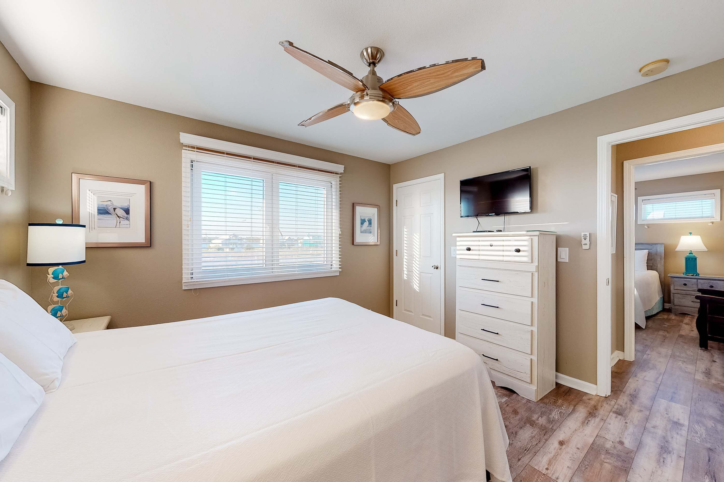 Une chambre à coucher comprenant un lit, un ventilateur de plafond, une télévision, une commode et une fenêtre avec des stores.