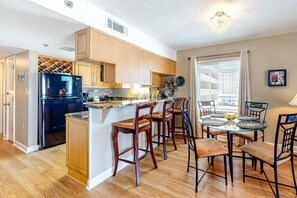 A kitchen with wooden cabinets, a black refrigerator, a glass island with bar stools, and a dining area with a glass table and chairs. - Gulf-front condo on 5th floor with amazing views, on-site pool & balcony (Gulf Shores)
