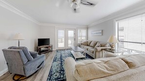 Ein Wohnzimmer mit grauem Sofa, beigem Sofa und einem Couchtisch. Ein Fernseher steht auf einem Schrank, eine Lampe und ein Deckenventilator sind ebenfalls vorhanden.