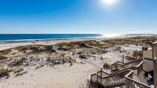 Ein Strand mit einer Holztreppe, die zu einem Gebäude führt, bewachsenen Dünen und dem Meer.