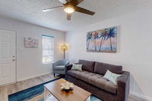 A living room with a ceiling fan, a sofa, a coffee table, a chair, a painting, and a window with blinds. - Comfortable single-level condo with WiFi, pool, & central AC (Gulf Shores)