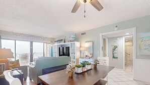 A spacious living area with a wooden dining table, a sofa, and a TV. There are potted plants on the table and a balcony with a railing. - Recently renovated 5th-floor oceanfront condo with pool, ocean view & WiFi (Gulf Shores)