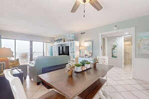 A spacious living area with a wooden dining table, a sofa, and a TV. There are potted plants on the table and a balcony with a railing. - Recently renovated 5th-floor oceanfront condo with pool, ocean view & WiFi (Gulf Shores)