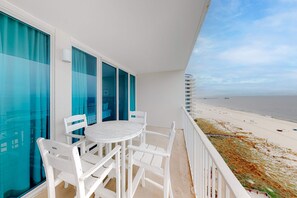 A balcony with a white table and chairs, a view of the beach, and a building in the background. - Polished, Gulf-Front Condo w/ Balcony, AC, WiFi & Shared Pools/Hot Tubs/Sauna (Gulf Shores)