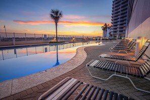 A poolside area with lounge chairs, a palm tree, and a high-rise building in the background. - Gulf-Front Condo w Gorgeous Views, Balcony & Shared Pools, Hot Tubs & Gym (Gulf Shores)