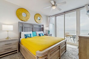 A bedroom with a bed, a nightstand, a lamp, a ceiling fan, and a balcony with a table and chairs. - Gulf-Front Condo w Great Balcony, Ocean View, AC & Shared Pools, Hot Tubs & Gym (Gulf Shores)