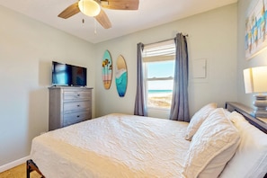 A bedroom with a bed, a TV, a ceiling fan, a window with curtains, and a surfboard art piece. - Beachfront condo with shared pool, outdoor shower, and beach access (Gulf Shores)