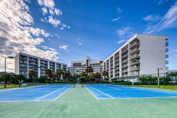 A tennis court with a net, surrounded by a fence, and adjacent to a multi-story building with balconies. - Completely renovated gulf view condo with pool, hot tub, tennis, and pickleball (Gulf Shores)