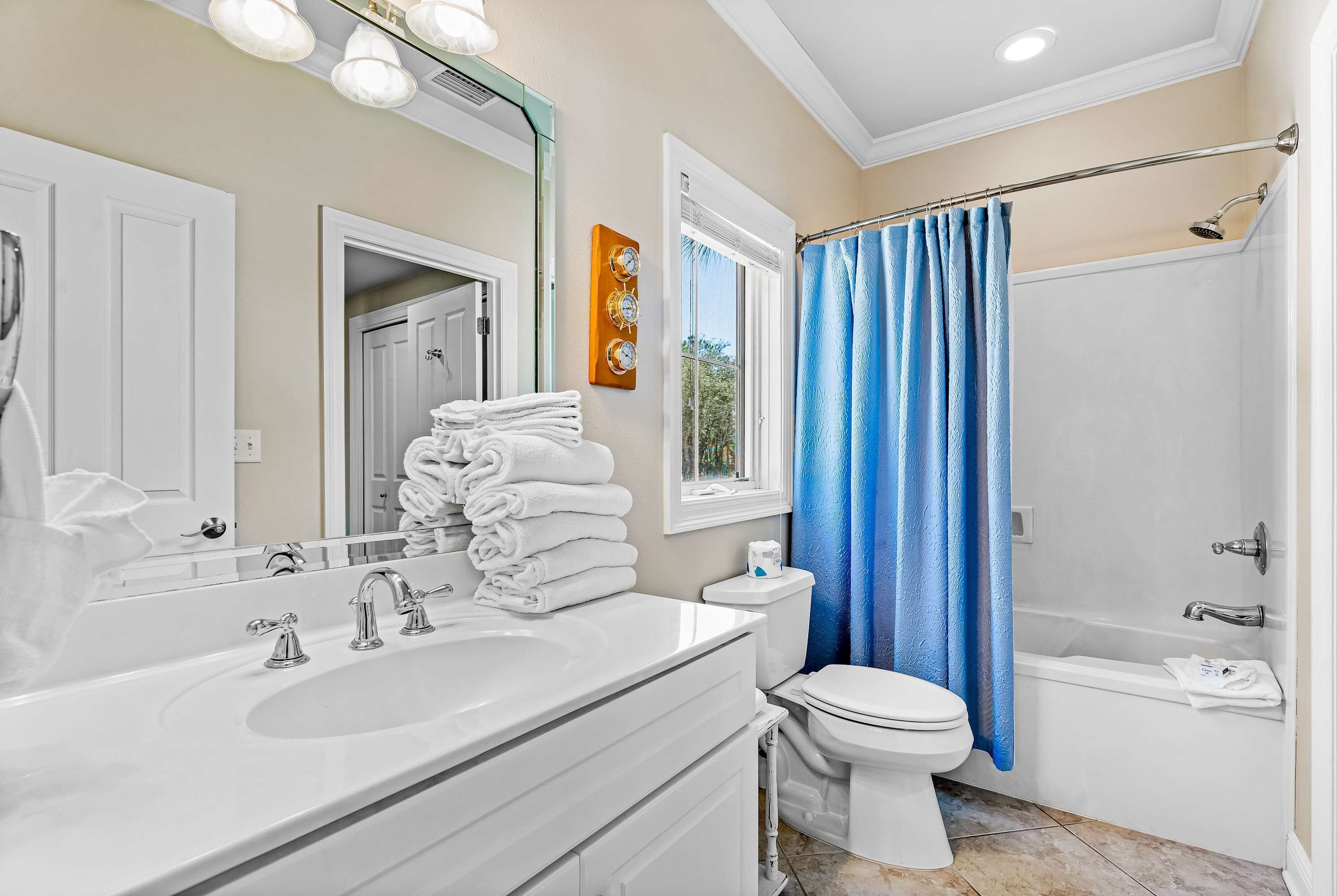 Une salle de bain avec un lavabo, des toilettes et une baignoire blancs, un rideau de douche bleu et une fenêtre donnant sur des arbres.