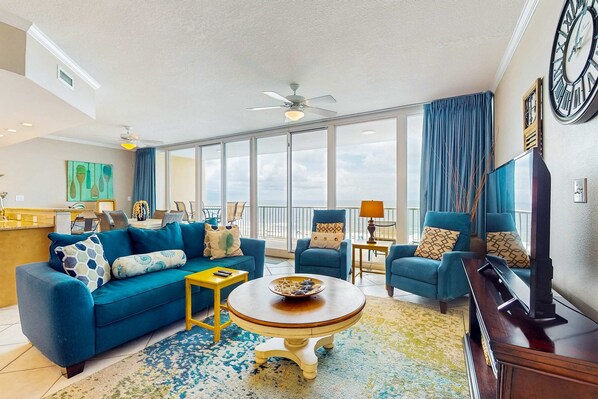 A living room with a blue sofa, a round coffee table, and a TV stand. - Gulf-view retreat with washer,dryer, AC, pool, sauna, & fitness center (Gulf Shores)