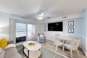 A modern living room with a ceiling fan, a large TV, a dining table with chairs, a sofa, and a balcony with a view. - 3rd-floor beachfront condo with pool, sauna, gym, hot tub, Gulf views, balcony (Gulf Shores)