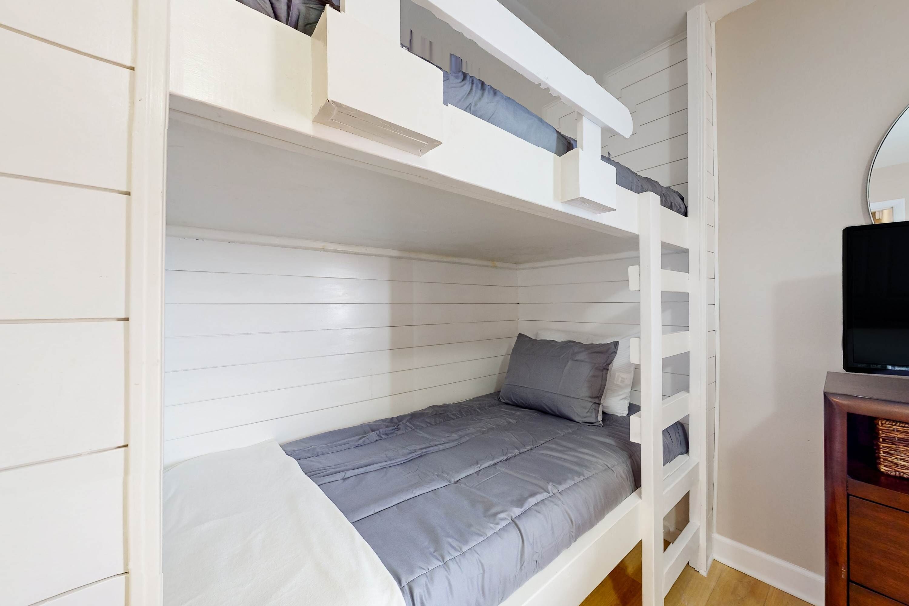 A bunk bed with a grey mattress and pillows, a wooden dresser with baskets, and a mirror on the wall.