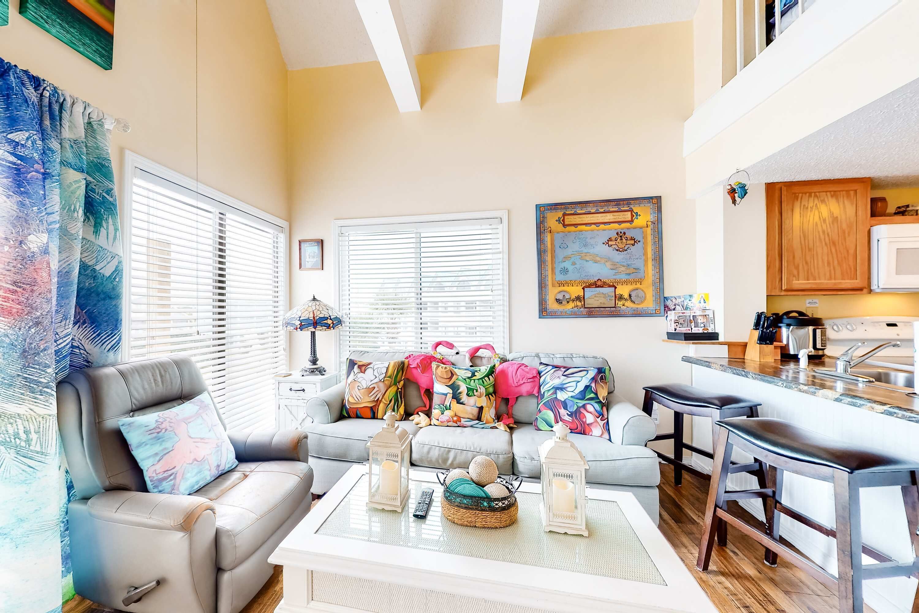 A living room with a sofa, a coffee table with decorative items, a kitchen area with a microwave, and a dining area with bar stools.