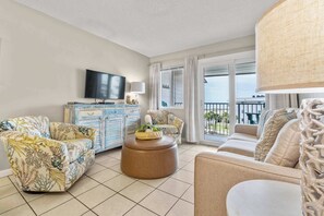 Ein Wohnzimmer mit Sofa, Sessel, Hocker, Fernseher und Blick ins Freie.