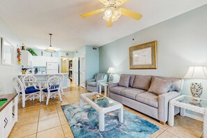 A living room with a sofa, coffee table, and chairs, a dining area with a table and chairs, and a kitchen in the background. - Unique dog-friendly Gulf-front condo with amazing views, pools, sauna, gym, & AC (Gulf Shores)