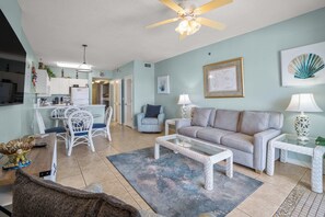A living room with a sofa, coffee table, and chairs, a dining area with a table and chairs, and a kitchen with cabinets and a refrigerator. - Unique dog-friendly Gulf-front condo with amazing views, pools, sauna, gym, & AC (Gulf Shores)
