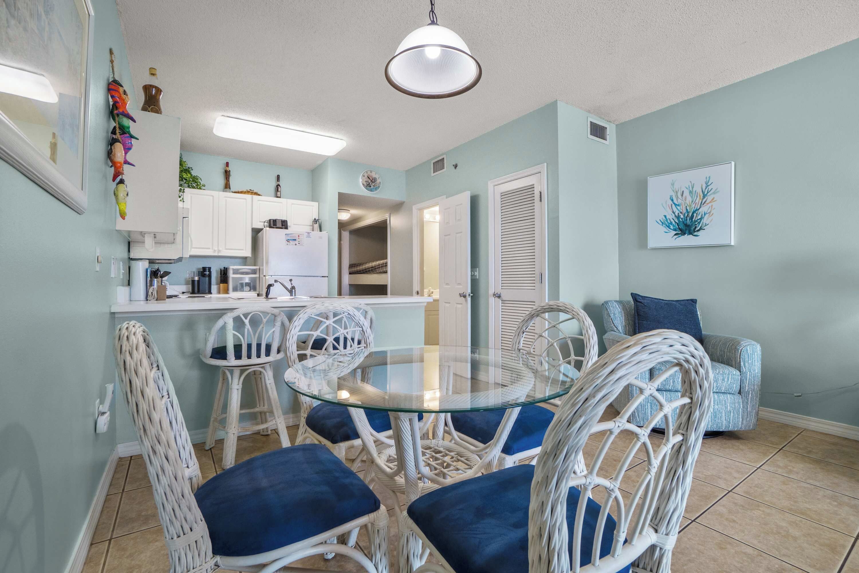 A compact living space with a dining area featuring a round glass table and white wicker chairs, a kitchenette, a living room with a sofa, and a wall-mounted clock.