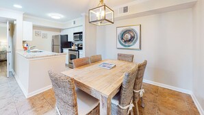 A dining area with a wooden table, wicker chairs, a hanging light fixture, a kitchen in the background, and a wall-mounted artwork. - Delightful coastal gem with pool, hot tub, private balcony, fast WiFi & AC (Gulf Shores)