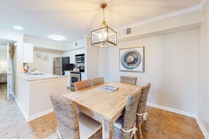 A dining area with a wooden table, wicker chairs, a hanging light fixture, a kitchen in the background, and a wall-mounted artwork. - Delightful coastal gem with pool, hot tub, private balcony, fast WiFi & AC (Gulf Shores)